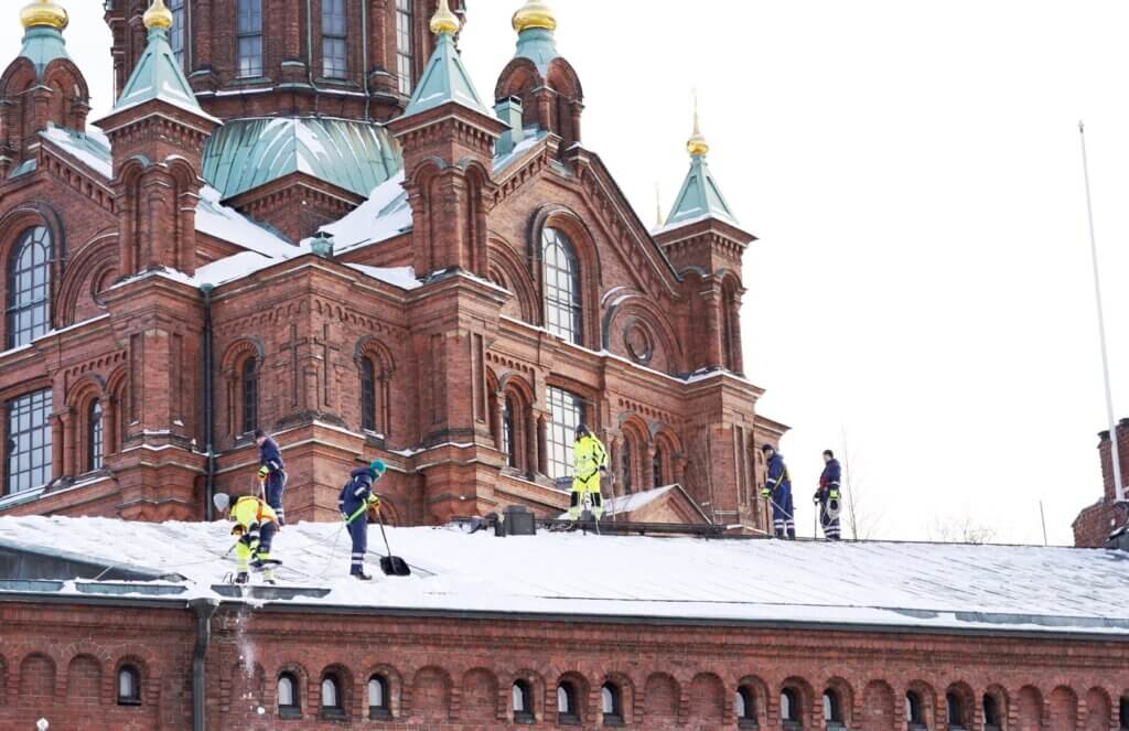 Snöskottning i Stockholm där vi ser sex stycken snöskottare skotta ner snö från ett tak på en tegelbyggnad